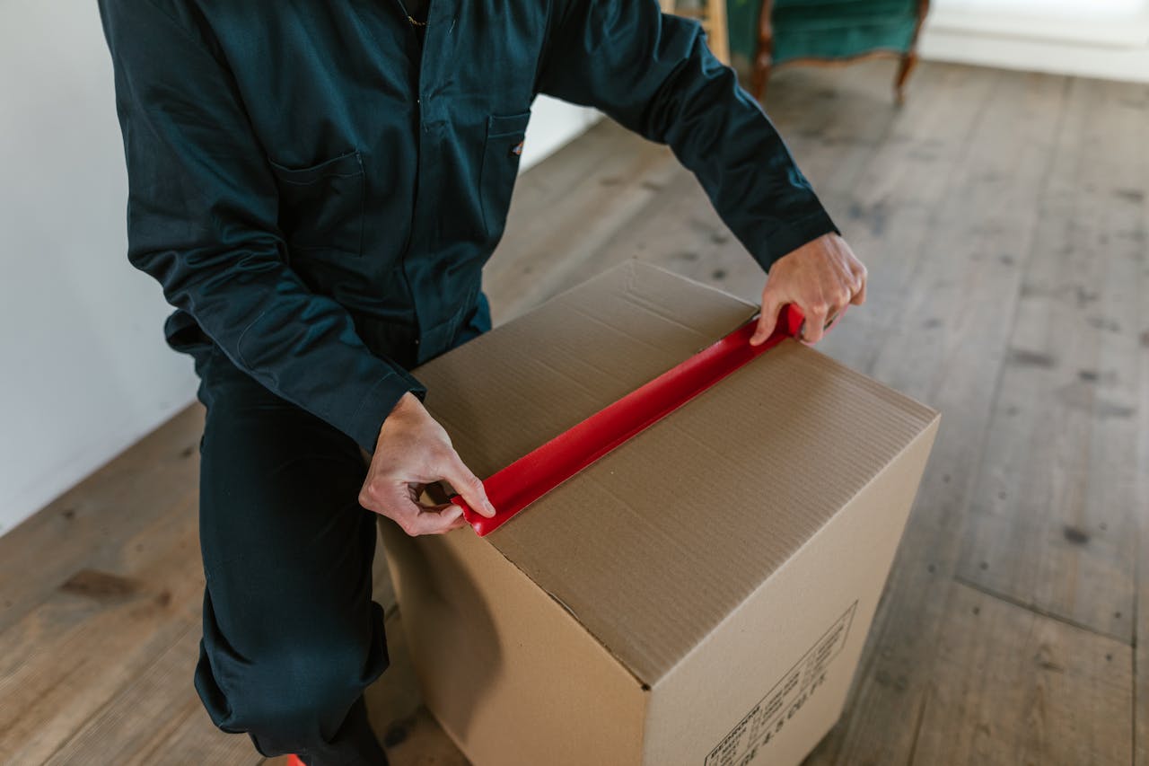 why-choose-us Male courier sealing a cardboard box with red tape, indoor wooden floor setting.
